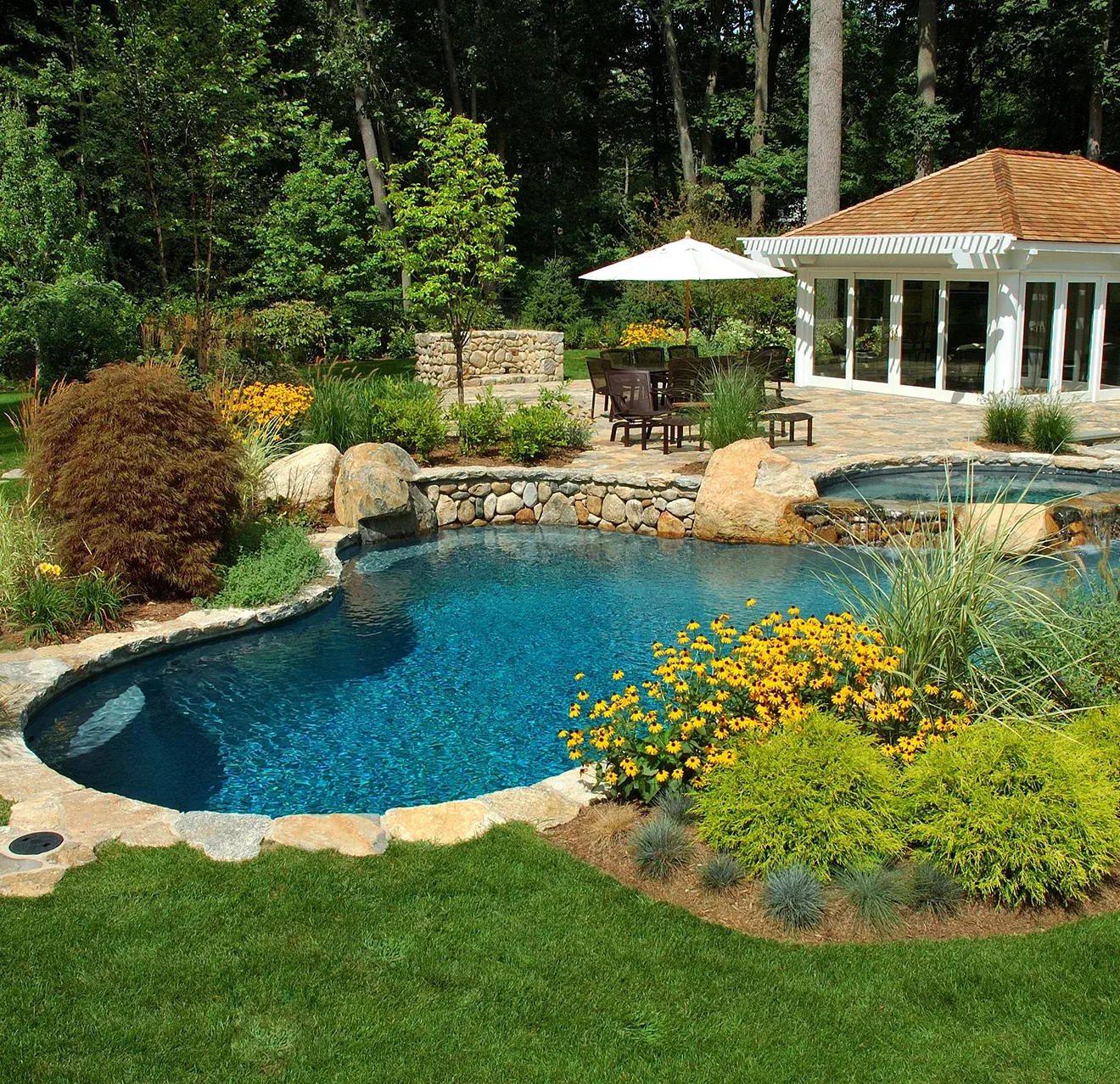 A inground pool with hot tub, patio, pool house, and lush landscaping.